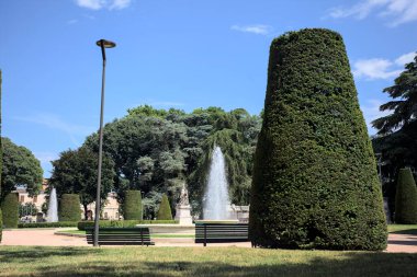 Çeşmelerle çevrili bir çeşme. Yanında ahşap banklar var. Güneşli bir günde bir parktaki çimlerin arkasından görülebiliyor.