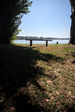 Gölün kıyısındaki çimenlikte bir bank. Ufukta karşı kıyıda parkı olan bir ağaç tarafından çerçevelenmiş.