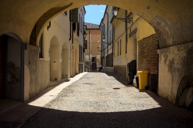 Güneş ışığındaki kaldırımlı cadde gölgedeki bir kemer yolu tarafından görünür ve çerçevelenir.