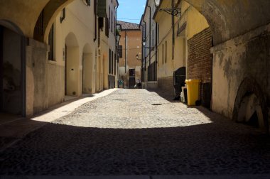 Güneş ışığındaki kaldırımlı cadde gölgedeki bir kemer yolu tarafından görünür ve çerçevelenir.