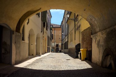 Güneş ışığındaki kaldırımlı cadde gölgedeki bir kemer yolu tarafından görünür ve çerçevelenir.