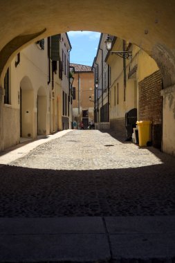 Güneş ışığındaki kaldırımlı cadde gölgedeki bir kemer yolu tarafından görünür ve çerçevelenir.