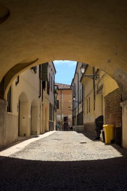Güneş ışığındaki kaldırımlı cadde gölgedeki bir kemer yolu tarafından görünür ve çerçevelenir.