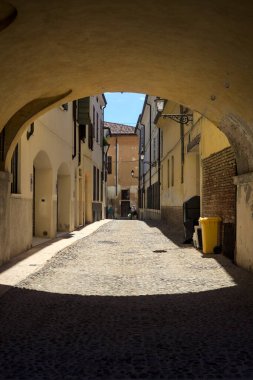 Güneş ışığındaki kaldırımlı cadde gölgedeki bir kemer yolu tarafından görünür ve çerçevelenir.