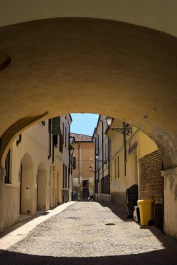 Güneş ışığındaki kaldırımlı cadde gölgedeki bir kemer yolu tarafından görünür ve çerçevelenir.