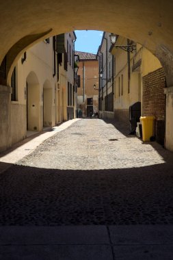 Güneş ışığındaki kaldırımlı cadde gölgedeki bir kemer yolu tarafından görünür ve çerçevelenir.