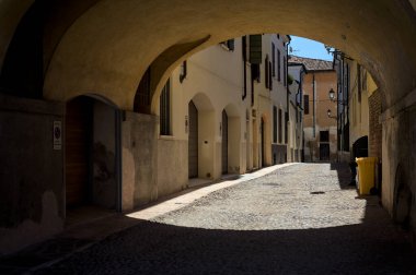 Güneş ışığındaki kaldırımlı cadde gölgedeki bir kemer yolu tarafından görünür ve çerçevelenir.