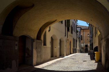 Güneş ışığındaki kaldırımlı cadde gölgedeki bir kemer yolu tarafından görünür ve çerçevelenir.