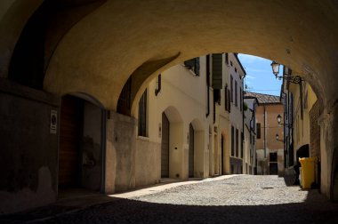 Güneş ışığındaki kaldırımlı cadde gölgedeki bir kemer yolu tarafından görünür ve çerçevelenir.