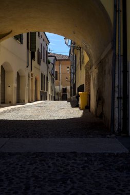 Güneş ışığındaki kaldırımlı cadde gölgedeki bir kemer yolu tarafından görünür ve çerçevelenir.