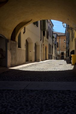 Güneş ışığındaki kaldırımlı cadde gölgedeki bir kemer yolu tarafından görünür ve çerçevelenir.