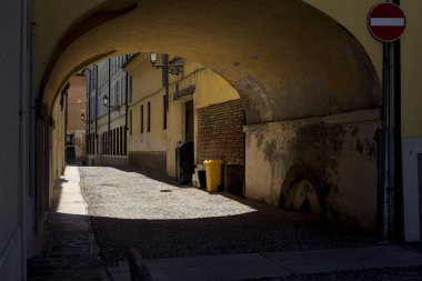 Güneş ışığındaki kaldırımlı cadde gölgedeki bir kemer yolu tarafından görünür ve çerçevelenir.