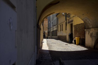Güneş ışığındaki kaldırımlı cadde gölgedeki bir kemer yolu tarafından görünür ve çerçevelenir.