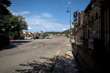 Verona, İtalya - 30 Temmuz 2025 - Nehrin yanında ve parkın kenarında yoldan geçen arabaların görüldüğü bir cadde