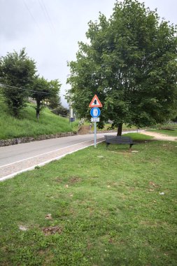 Bulutlu bir günde, bir İtalyan köyünün çimlerinden görünen bir ağaç ve bir tabela ile dağ yolu.