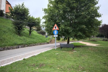 Bulutlu bir günde, bir İtalyan köyünün çimlerinden görünen bir ağaç ve bir tabela ile dağ yolu.