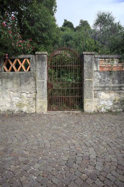Paslı bir kapı eskimiş bir beton duvarın içinde. Arkasında da bulutlu bir günde asfalt bir patikanın kenarında bir bahçe var.