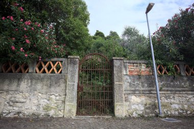 Paslı bir kapı eskimiş bir beton duvarın içinde. Arkasında da bulutlu bir günde asfalt bir patikanın kenarında bir bahçe var.