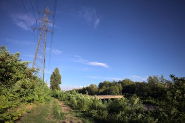 Yanı başında direği olan bitkilerle çevrili bir patika ve gün batımında İtalya 'nın kırsal kesimindeki bir eğlence kanalının kenarında bir elektrik hattı var.