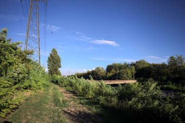 Yanı başında direği olan bitkilerle çevrili bir patika ve gün batımında İtalya 'nın kırsal kesimindeki bir eğlence kanalının kenarında bir elektrik hattı var.