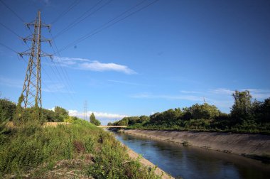 Bir patikanın yanında tarla sürmüş ve üzerinden elektrik hattı geçen saptırıcı bir kanal ve gün batımında İtalya kırsalında direği olan bir kanal.