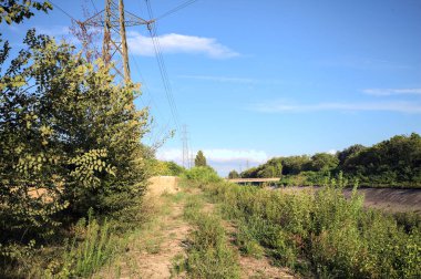 Yanı başında direği olan bitkilerle çevrili bir patika ve gün batımında İtalya 'nın kırsal kesimindeki bir eğlence kanalının kenarında bir elektrik hattı var.