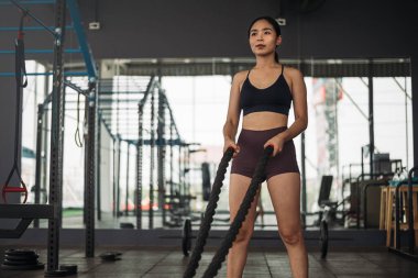Güçlü Asyalı kadın Crossfit spor salonunda savaş halatıyla egzersiz yapıyor. Spor kıyafetleri giyen atlet kadın gri spor salonunda ağırlık ve halterli. Sağlıklı yaşam tarzı.