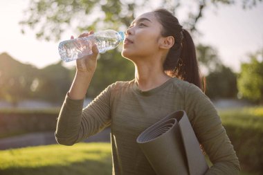 Spor giyimli, su içen Asyalı bir kadın ve güneş ışığında parkta egzersiz yapmak için yoga minderi tutuyor. Sağlık ve Sağlık Faaliyetleri.