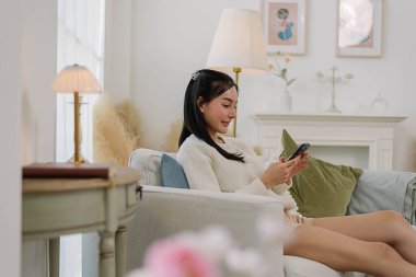 Konforlu oturma odasında akıllı telefon kullanan Asyalı bir kadın, rahat bir an geçirip şık dekore edilmiş oturma odasında akıllı telefonuyla sohbet etmekten hoşlanıyor. Yaşam tarzı ve günlük aktiviteler.