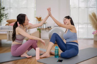 Aktif kıyafetli iki Asyalı kadın, rahat bir odada başarılı bir çalışma yaptıktan sonra yoga minderlerine oturup beşlik çakıyorlar. Sağlık ve sağlık hizmetleri faaliyetleri.
