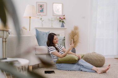 Asyalı bir kadın rahat oturma odasında selfie çekiyor, genç bir bayan şık dekore edilmiş oturma odasında akıllı telefonuyla rahatça fotoğraf çekmekten hoşlanıyor. Yaşam tarzı ve günlük aktiviteler.