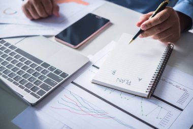 Dağınık masada oturan bir adam, A ya da B planı yapıyor, not defterine planlar yazıyor.