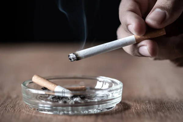 男人手拿着燃烧的香烟在木桌上的烟灰缸上 免版税图库照片