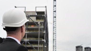 İnşaatla ilgili bazı detaylara el ile işaret eden mimar, arka plandaki inşaat alanlarında vinç ve iş elbiseleriyle tamamlanmamış bina mimarı ve sokakta beyaz kask hareketi