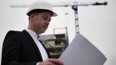 Mimar, elinde tuttuğu kağıt çizimleriyle ilgili ayrıntıları bir inşaat vincinin arka planında ve tamamlanmamış bir binada inceliyor. İş kıyafetli bir mimar ve sokakta beyaz kask takıyor.