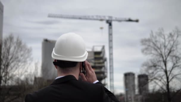 一位身穿西装 头戴白色头盔的建筑师 手里拿着手机 与人交谈 在前面的背景下 他领导着建筑和建筑用起重机的建设 — 图库视频影像