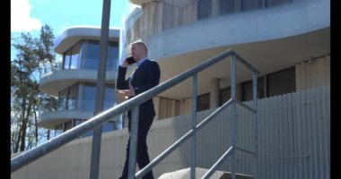 İş kıyafetleri içinde şık giyinen bir işadamı, şehir caddesinde bir ofis kulesiyle dikilirken telefonuyla konuşur. İş adamlarının etkileşimi acil iş iletişimi anlamına gelir.