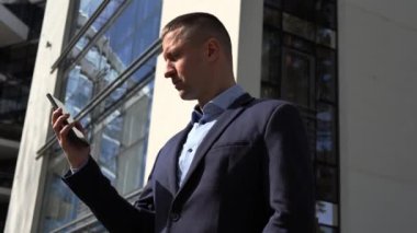 İş adamı telefonu hızla ofis binasının arka planına kaldırır. İşadamı ciddi bir diyalog içinde. Yoğunluk işaretlendi. İş adamlarının stratejik sohbetleri dikkatinin ve uzmanlığının altını çiziyor.