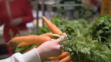 Kadın havucu dikkatlice seçiyor, elinde yeşillikle havuç tutuyor, markette havuçla sağlıklı seçimler yapıyor. Tazelik ve organik ürün seçiminin vurgulanması Ekolojik gıda kavramı