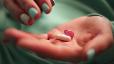 Bu video, kadın ellerini yakından gösteriyor, parmaklar nazikçe palmiye yeşili renk giysilerden ve tırnaklardan haplar alıyor çarpıcı bir kontrastı, hapları vurguluyor ve ilaçların dikkatle kullanılmasını sağlıyor..