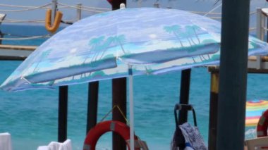 Mavi şemsiye kumsalda duruyor, rüzgarda hafifçe kıvrılıyor, dingin manzara şemsiyesine renk katıyor arka plandaki denizle çok güzel zıtlaşıyor ve sahnenin odak noktasını şemsiye haline getiriyor..