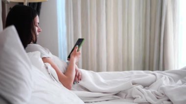 Genç esmer kadın, beyaz battaniyenin altında yatağında, telefonuna ilgi ile bakıyor genç esmer kadın barışçıl ifade onun sakin sabah rutinini vurguluyor.