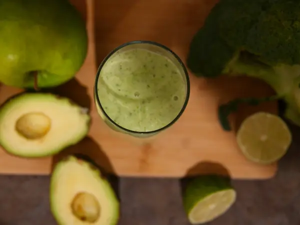 stock image Green smoothie in glass surrounded by avocado, lime, broccoli, apple, cucumber. Perfect smoothie for health, detox, and nutrition content. fresh ingredients and benefits of this vibrant smoothie.