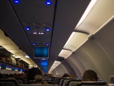 Yolcuların oturduğu dolu bir uçak kabini, yukarıda bagaj rafları ve gökyüzü manzaralı uçak kabini sunan pencereler uçuş beklentisi ve uğultusuyla dolu. Uçak kabini yaşamının özü.