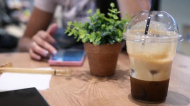 Masadaki plastik bardakta soğuk kahve, arka plandaki kadın telefona bakarken soğuk kahve zengin rengiyle göze çarpıyor, bitki ise yeşile dokunuyor. Soğuk kahve tazeliyorum. 