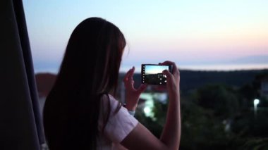 Genç bir kadın telefonda gün batımını çekiyor, gün batımının gökyüzünü boyarken canlı renkleri yakalıyor. Nefes kesici günbatımı yüzünü yansıtıyor, güzel ve huzurlu bir an yaratıyor..