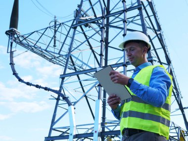 Beyaz bir miğfer, yansıtıcı yelek ve tabletle bir enerji mühendisi elektrik kulesi ve kabloları inceliyor. Enerji mühendisi verimli güç sistemlerini korumaya odaklanıyor.