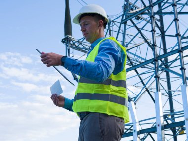 An energy engineer with a tablet, white helmet, and reflective vest surveys an electric tower and wires. This energy engineer ensures proper maintenance and safety of power infrastructure. clipart