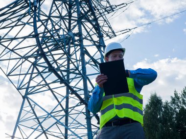 Beyaz miğferli ve yansıtıcı yelekli bir enerji mühendisi elinde tabletle bir elektrik kulesini ve kabloları inceliyor. Bu enerji mühendisi kendini güvenli ve verimli güç dağılımını sağlamaya adamıştır..