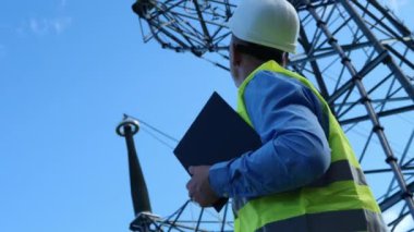 Beyaz miğferi, yansıtıcı yeleği ve tableti olan bir enerji mühendisi bir elektrik kulesini ve kabloları inceliyor. Bu enerji mühendisi kendini güvenli ve verimli güç sistemi yönetimine adamıştır..